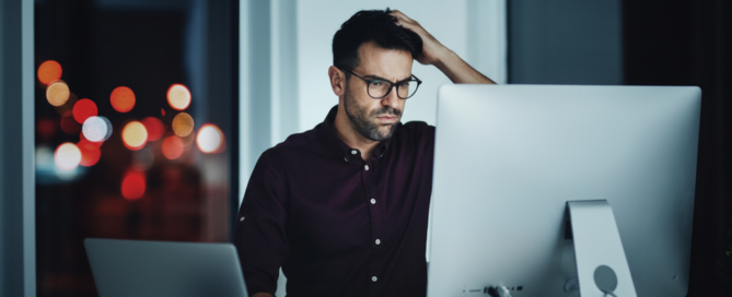 A man at a desk after dark, hand on his head, frowning at a monitor with city lights blurred behind him.